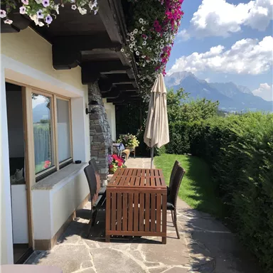 Ein einladender Außenbereich mit einem Tisch und Stühlen unter einem Sonnenschirm. Über dem Tisch blühen bunte Blumen und die Berge sind im Hintergrund sichtbar.