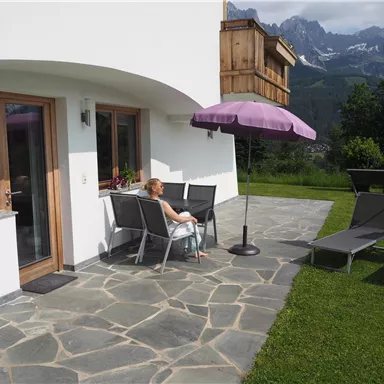 Eine Frau sitzt auf einer Terrasse vor einem modernen Haus. Im Hintergrund sind Berge und ein klarer Himmel zu sehen.