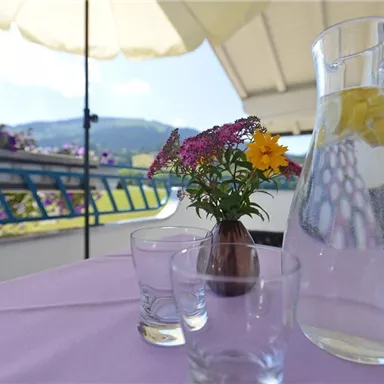 A beautiful table on a balcony with a glass carafe and two drinking glasses. A vase with colorful flowers and a sunshade complete the cozy atmosphere.