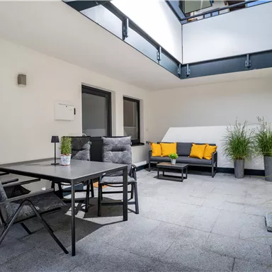 A modern courtyard with a cozy seating area and a table. The plants and the sunshade create an inviting atmosphere.