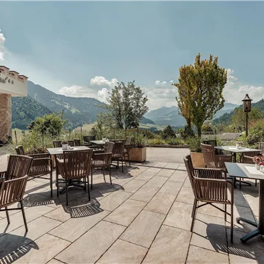 Eine sonnige Terrasse mit Holzstühlen und Tischen. Im Hintergrund sind Berge und ein klarer Himmel zu sehen.