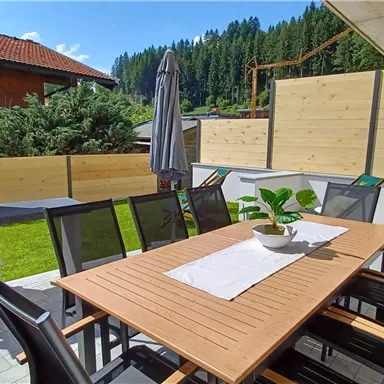 Eine moderne Terrasse mit einem großen Tisch und Stühlen. Im Hintergrund befindet sich ein Grill und eine gepflegte Gartenlandschaft.