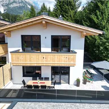 Ein modernes Haus mit einem Balkon und einer Terrasse. Umgeben von Bäumen und Bergen bietet es eine ruhige Atmosphäre.