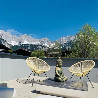 Eine sonnige Dachterrasse mit zwei Stühlen und einer Buddha-Statue. Im Hintergrund erheben sich beeindruckende Berge unter einem klaren blauen Himmel.