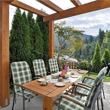 A beautiful outdoor dining area, surrounded by green plants. The table is set festively and offers a view of the mountains.