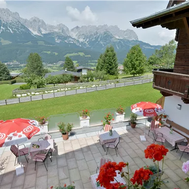 A beautiful terrace with seating and red sun umbrellas. In the background, impressive mountains and a green landscape can be seen.