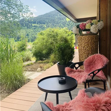 Ein gemütlicher Balkon mit zwei schwarzen Stühlen und einem kleinen Tisch. Im Hintergrund sind grüne Berge und dekorative Pflanzen zu sehen.