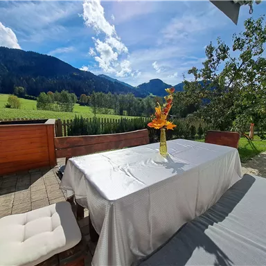 Ein schöner Tisch im Freien mit einem Blumenstrauß, umgeben von einer grünen Wiese und Bergen im Hintergrund. Der Himmel ist blau mit einigen Wolken.