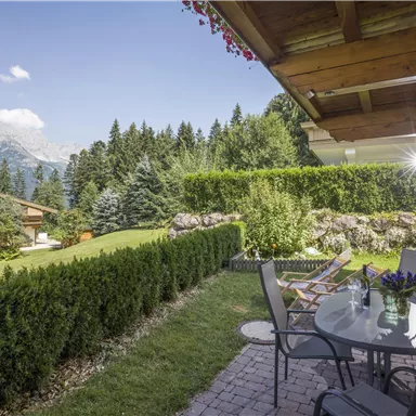 Ein einladender Garten mit gepflegten Hecken und Blick auf die Berge. Auf der Terrasse stehen Tische und Stühle, ideal für entspannte Stunden im Freien.