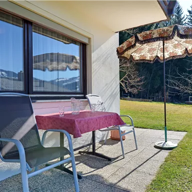 A terrace with a table and chairs as well as a sunshade. In the background, trees and another building are visible.