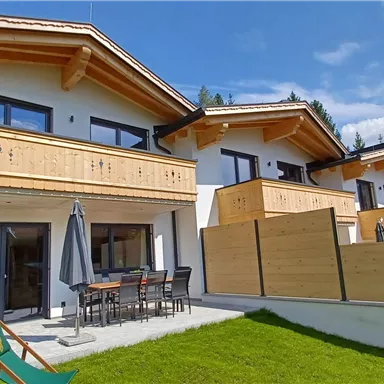 Ein modernes Haus mit Holzverkleidung und Balkon. Der Garten ist grün und einladend, umgeben von Bergen.