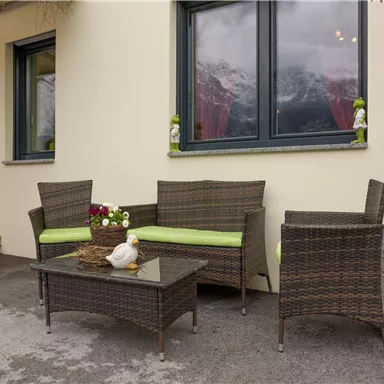 Ein gemütlicher Außenbereich mit einem Tisch und vier Stühlen aus Rattan. Die Sitze sind mit grünen Kissen ausgestattet und es gibt Blumendekorationen im Hintergrund.