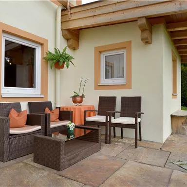 Eine gemütliche Terasse mit einem Rattanmöbelset und einem Tisch. Im Hintergrund sind ein Garten und ein Holzfass zu sehen.