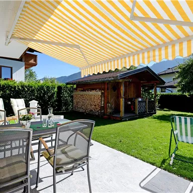 Eine gemütliche Terrasse mit einem Sonnenblenden und Gartenmöbeln. Im Hintergrund sind ein Garten und ein Holzschuppen sichtbar.