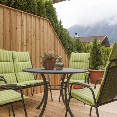 Ein schöner Außenbereich mit einem Tisch und vier Stühlen in grün gestreiftem Design. Im Hintergrund sind grüne Pflanzen und eine Berglandschaft sichtbar.