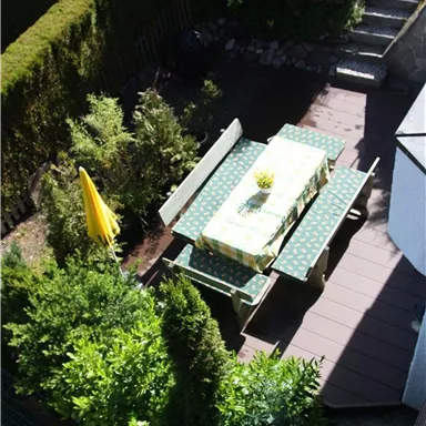 Ein gemütlicher Gartentisch mit grüner Tischdecke und Stühlen steht umgeben von Büschen. Im Hintergrund sind einige Treppen und eine gelbe Sonnenschirm zu sehen.