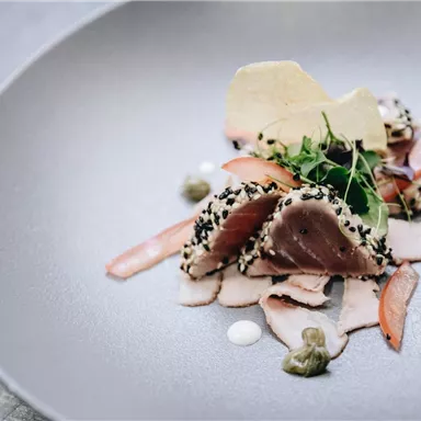 An artistic dish with sea fish, enveloped in black sesame. The meal is garnished with fresh herbs and sauces on an elegant plate.