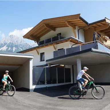 A modern house with wooden decor and a beautiful mountain view. Two cyclists are riding on the access road.
