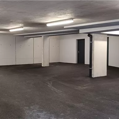 Ein leerer Raum mit Betonboden und weißen Wänden. Es gibt mehrere Türen und Deckenlichter.