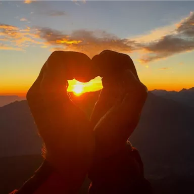 Zwei Hände formen ein Herz vor einem Sonnenuntergang über den Bergen. Der Himmel erstrahlt in warmen Farben.