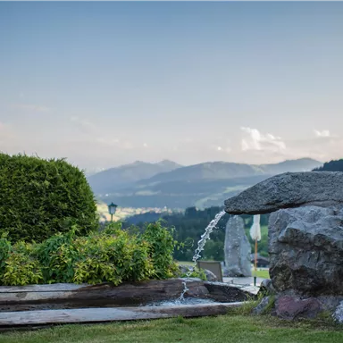 Eine ruhige Landschaft mit einem Wasserfall, der über einen Steinhaufen fließt. Im Hintergrund sind sanfte Hügel und Berge sichtbar.