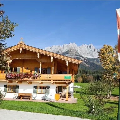 A traditional alpine wooden house surrounded by meadows and mountains. The flag in the foreground indicates the region.