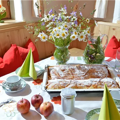 An inviting table with a freshly baked apple strudel, apples, and tea cups. A vase with colorful flowers and green napkins complete the cozy atmosphere.
