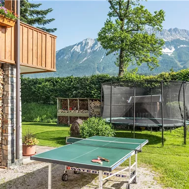 Ein schöner Garten mit einem Tischtennistisch und einem Trampolin. Im Hintergrund sind Berge und ein klarer blauer Himmel zu sehen.