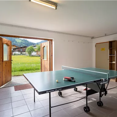 Ein heller Raum mit einem Tischtennistisch und einer offenen Tür, die auf eine grüne Wiese blickt. Im Hintergrund steht ein Kicker und an der Wand sind Garderobenhaken angebracht.