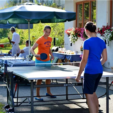 Two girls are playing table tennis in the garden. In the background, cheerful guests and blooming plants can be seen.