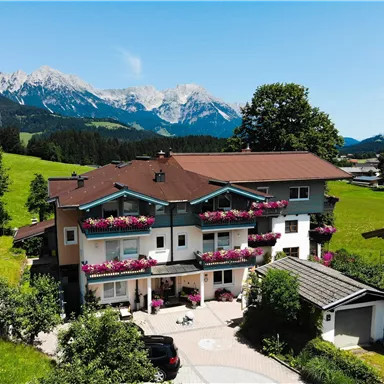 Ein malerisches Haus in der Natur mit Blumenkästen an den Balkonen. Im Hintergrund sind die majestätischen Berge und ein blauer Himmel zu sehen.