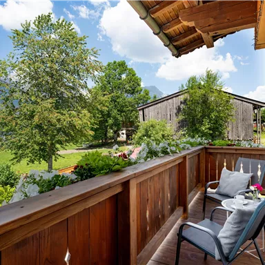 A balcony with two chairs and a table, surrounded by plants. In the background, trees and mountains can be seen under a blue sky.