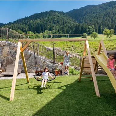 A playground with a swing and a slide. In the background, green meadows and hills can be seen.