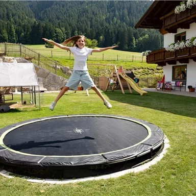 A girl happily jumps on a trampoline in a green garden. In the background, a beautiful house and a mountainous landscape can be seen.