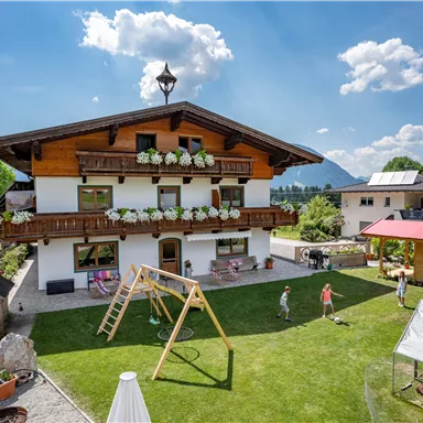 A beautiful country house with a large garden and playground. In the background, mountains and a clear sky can be seen.