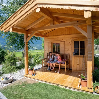 A cozy wooden terrace with two people sitting relaxed. In the background, a green landscape and mountains can be seen.