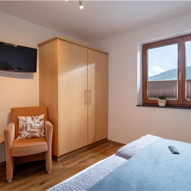 A modern bedroom with a double bed and a television on the wall. Large windows provide a view of the mountains and plenty of daylight.