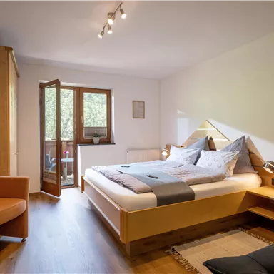 A bright and modern bedroom with a double bed and a cozy armchair. A balcony with a window provides fresh air and daylight.