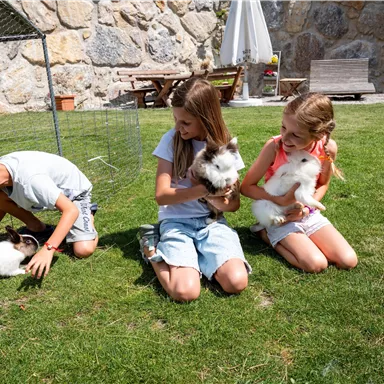 Three children are playing outdoors with rabbits on a green meadow. The sun is shining, and in the background, some seating areas can be seen.