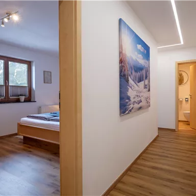A bright hallway with a wooden floor and modern design. On the left side, there is a bedroom and a piece of art on the wall.