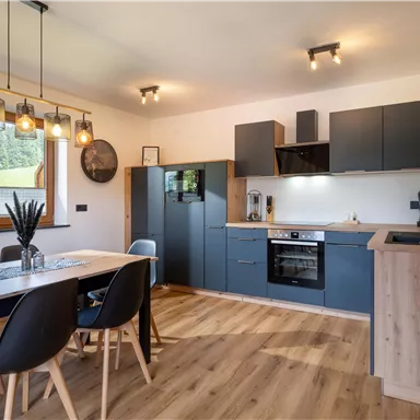 A modern kitchen with wood-colored flooring and elegant dark cabinets. A large dining table with chairs is in the room, which has plenty of natural light.
