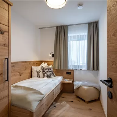 Ein gemütliches Zimmer mit Holzmöbeln und einem Fenster mit Vorhängen. Es gibt ein Einzelbett und einen kleinen Sitzbereich.