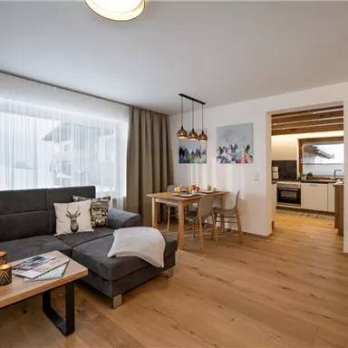 Eine moderne und helle Wohnküche mit einem gemütlichen Sofa und einem Esstisch. Die großen Fenster sorgen für viel Tageslicht und eine einladende Atmosphäre.