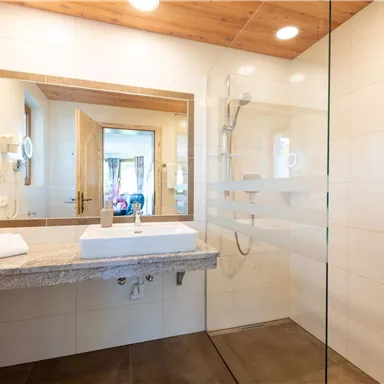 Ein modernes Badezimmer mit einer großzügigen Dusche und einem Waschbecken auf einer Steinablage. Große Fliesen und Holzelemente verleihen dem Raum eine helle, einladende Atmosphäre.