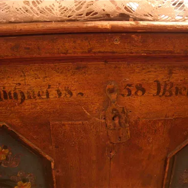 An antique piece of wood with intricate painting and detailed writing. It displays a blend of floral patterns and historical embellishments.