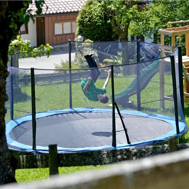 A child is jumping on a trampoline in the garden. In the background, a playground and trees can be seen.