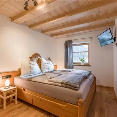 A cozy bedroom with wooden furniture and a large window. The room is bright and modernly furnished, with a small table and a TV on the wall.