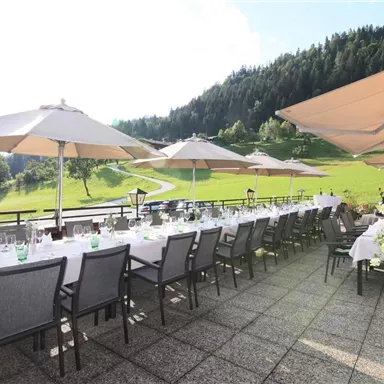 An inviting terrace with long covered tables and sun umbrellas. In the background, a green meadow and wooded hills stretch out.