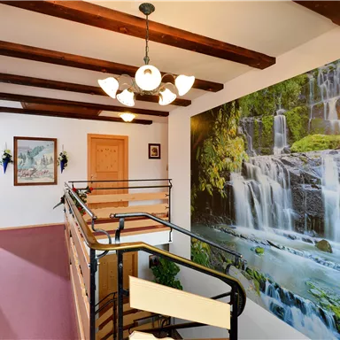 Ein helles Treppenhaus mit Holzdeckenbalken und einer großen Fototapete eines Wasserfalls. Der Raum ist freundlich und einladend gestaltet.