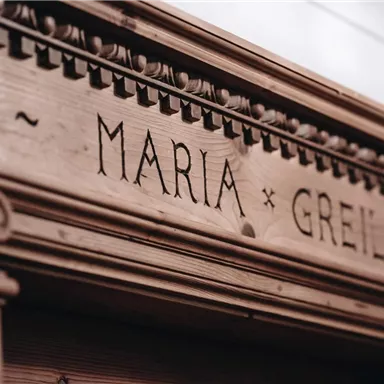 An artistically carved wooden frame with the engraved name "Maria Greil". The structure displays fine details and gives the room a nostalgic atmosphere.